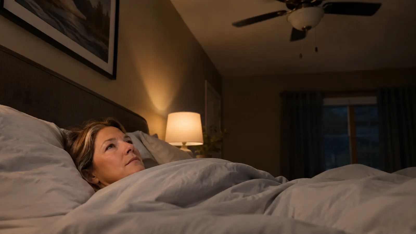 A woman in her fifties lying on a queen bed with a sheet pulled to her chin, ceiling fan visible above, dim evening light