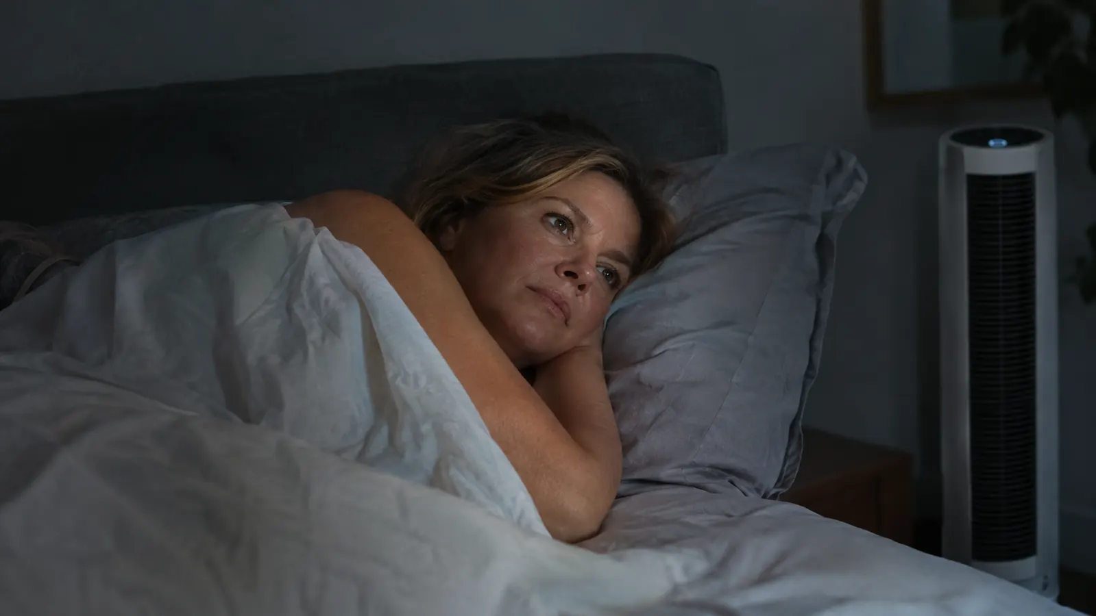 A woman in her fifties lying on her side under a light sheet with a small tower fan beside the bed