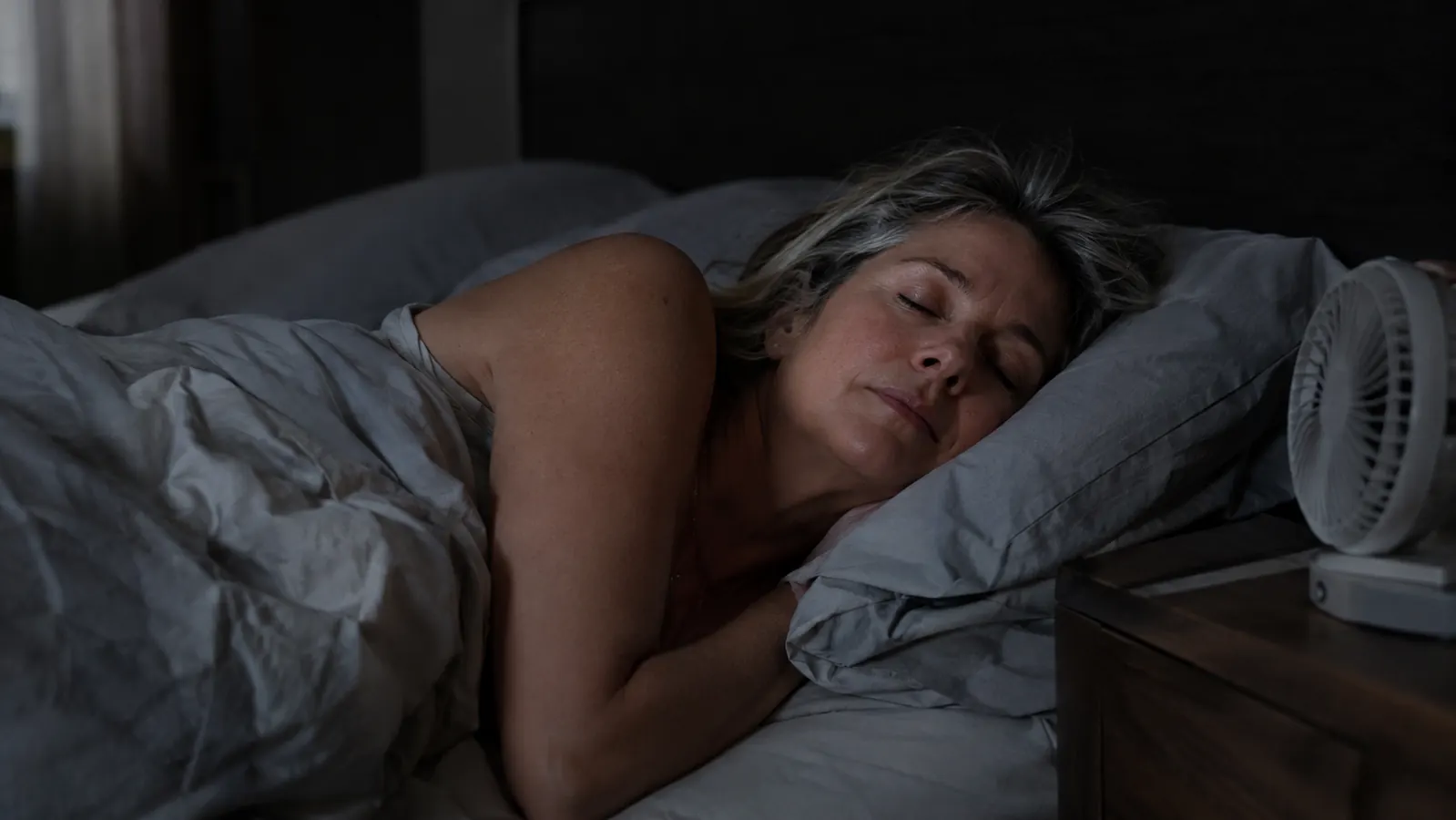 A woman in her fifties asleep under a light sheet in a dark bedroom, one arm outside the covers