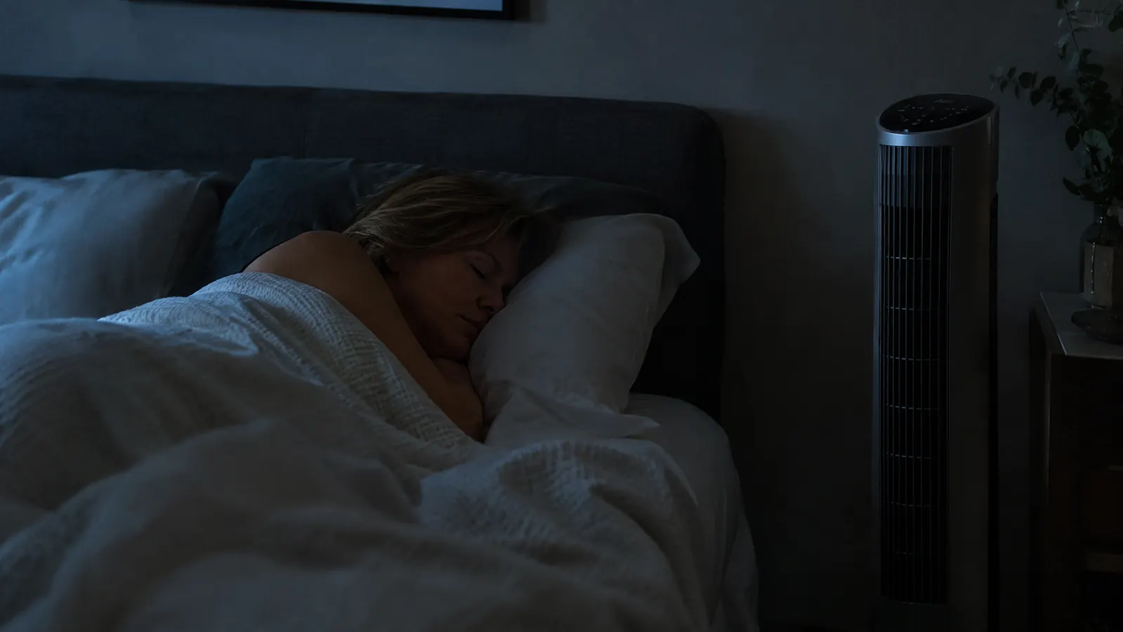 A woman in her fifties asleep under a light blanket, a fan standing beside the bed